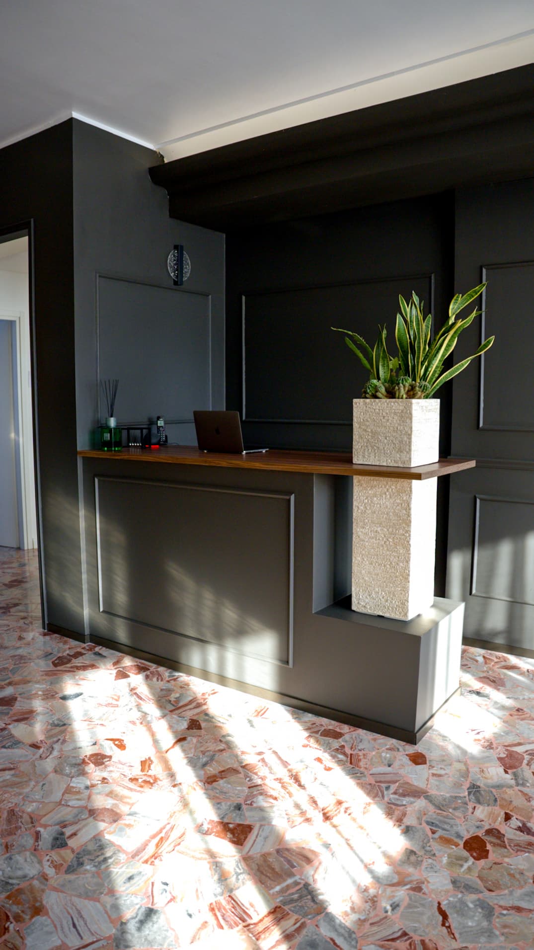 Reception con boiserie antracite, piano in legno e fioriera in travertino illuminata dal sole